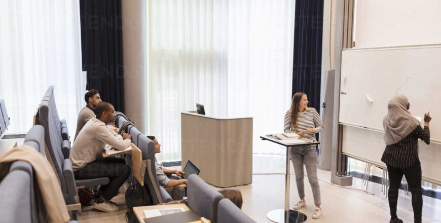 Students looking at friend writing on whiteboard during rehearsal in lecture hall